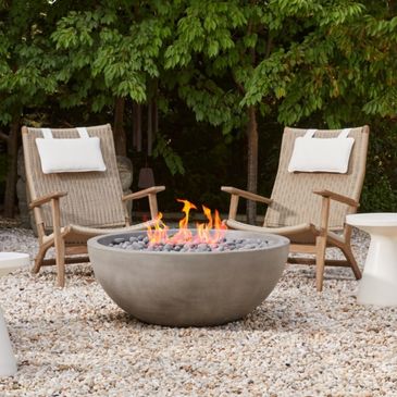 Outdoor fire pit with two cushioned chairs on gravel under green trees.