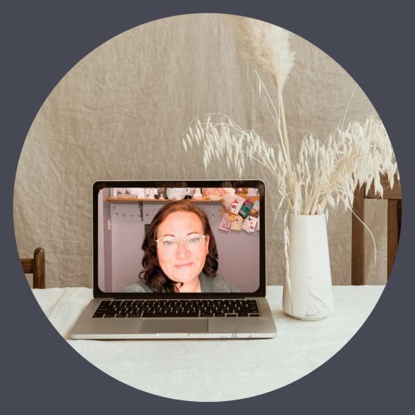 Laptop on table with a woman on screen and a vase of dried plants.