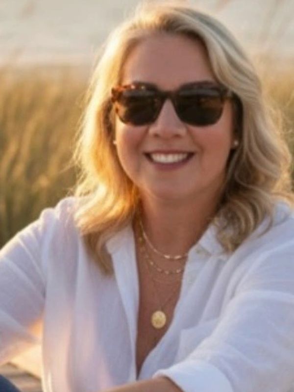 Smiling woman with blonde hair wearing sunglasses and a white shirt outdoors.