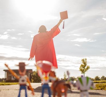 Graduate in red gown holding cap celebrating outdoors.