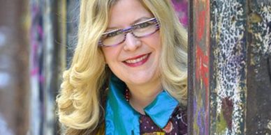 Smiling woman with blonde hair and glasses peeking from behind a colorful graffiti wall.