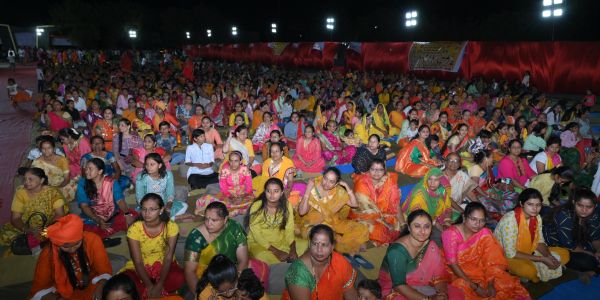 Large crowd of people sitting on the ground at a night event, dressed in colorful traditional attire.