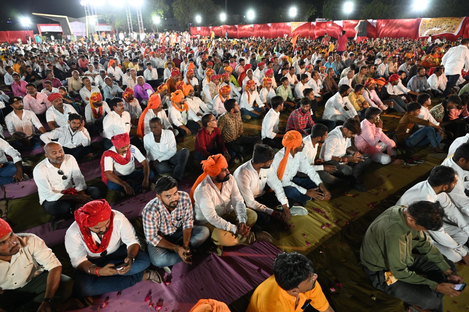 A large crowd of men sitting on the ground, many wearing orange turbans and white shirts.