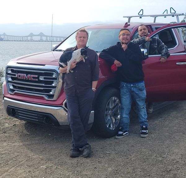 Three men posing by a red GMC truck near a waterfront, one holding a small dog.