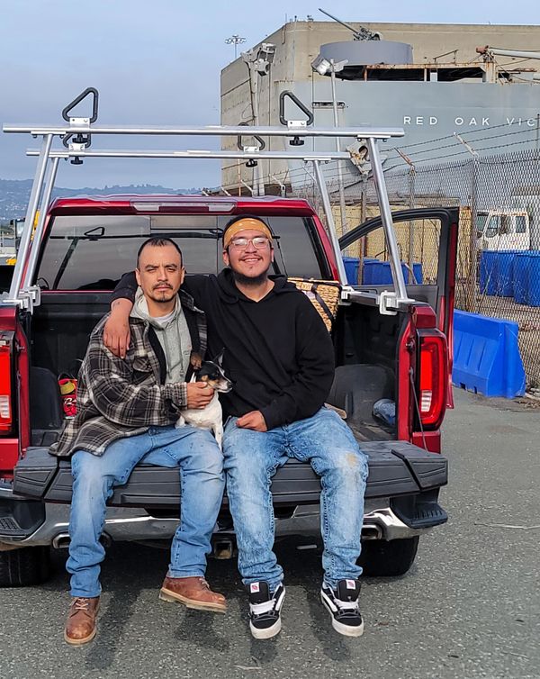 Two men and a dog sitting on the tailgate of a red pickup truck outdoors.