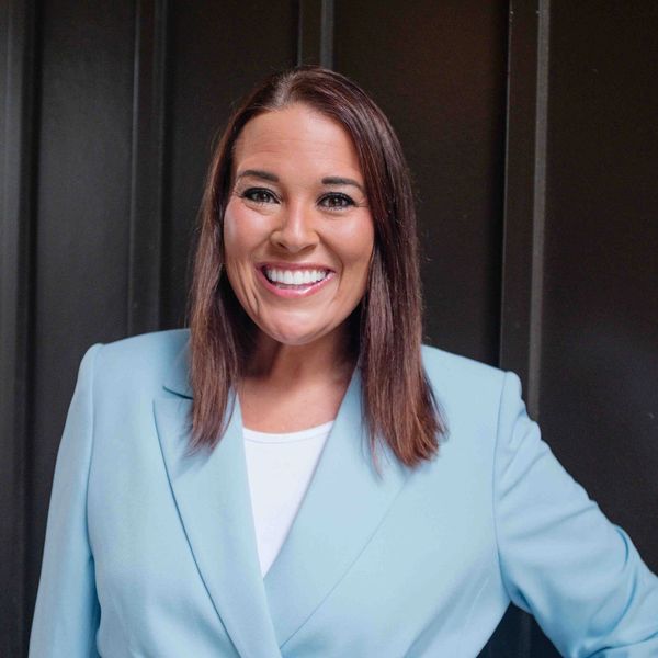 Smiling woman in a light blue blazer against a dark background.