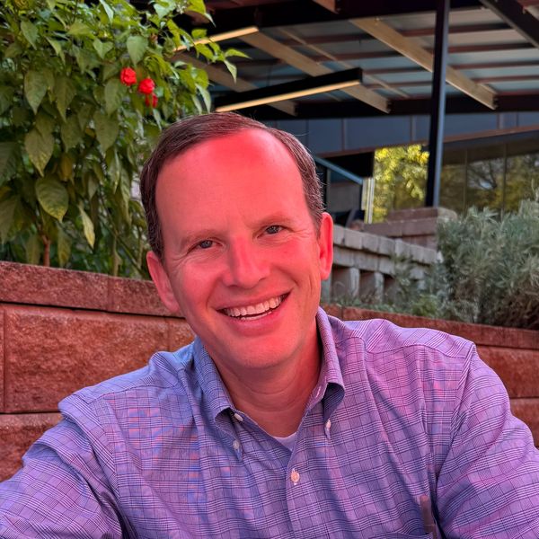 Smiling man in a checkered shirt sitting outdoors near plants and a brick wall.