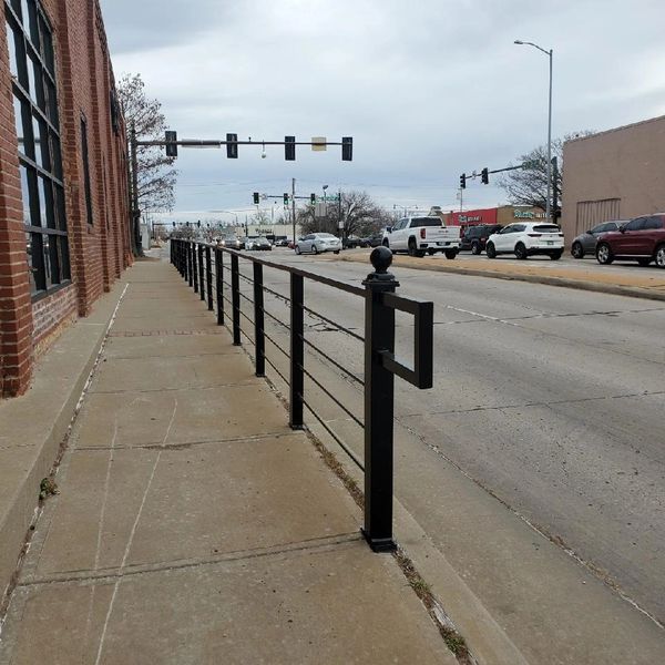 Sidewalk with black metal railing alongside a brick building near a busy street.
