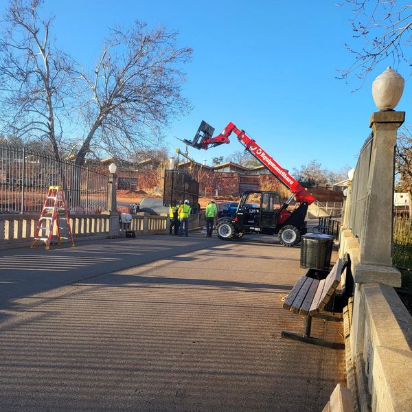 Workers using a red forklift on a sunny pathway near benches and a fence.