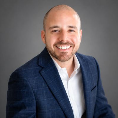Smiling man in a blue checkered blazer and white shirt against a gray background.
