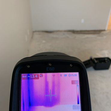 Thermal imaging device shows temperature variations on a wall and floor in an unfinished room.
