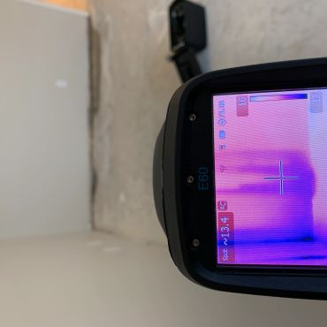 Thermal imaging device shows temperature variations on a wall and floor in an unfinished room.