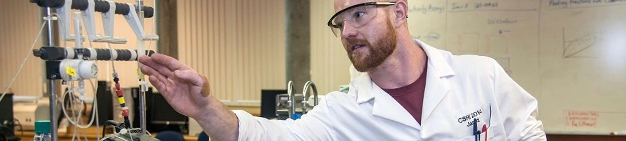 Scientist in lab coat and safety glasses reaching towards lab equipment.