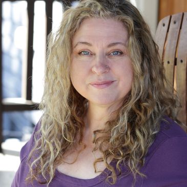 Smiling woman with curly blonde hair in a purple top sitting on a wooden chair.