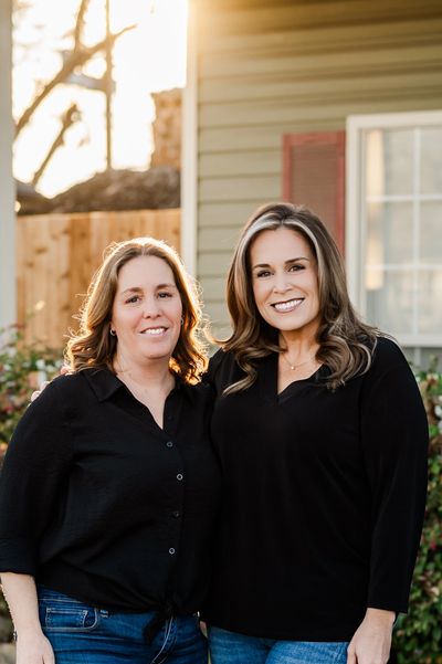 Two women in black tops and jeans smiling outside a house.