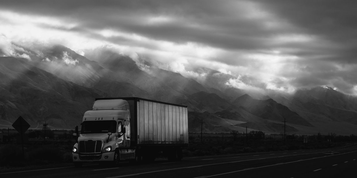 Black and white photo of a freight truck driving in front of mountains on a cloudy day.