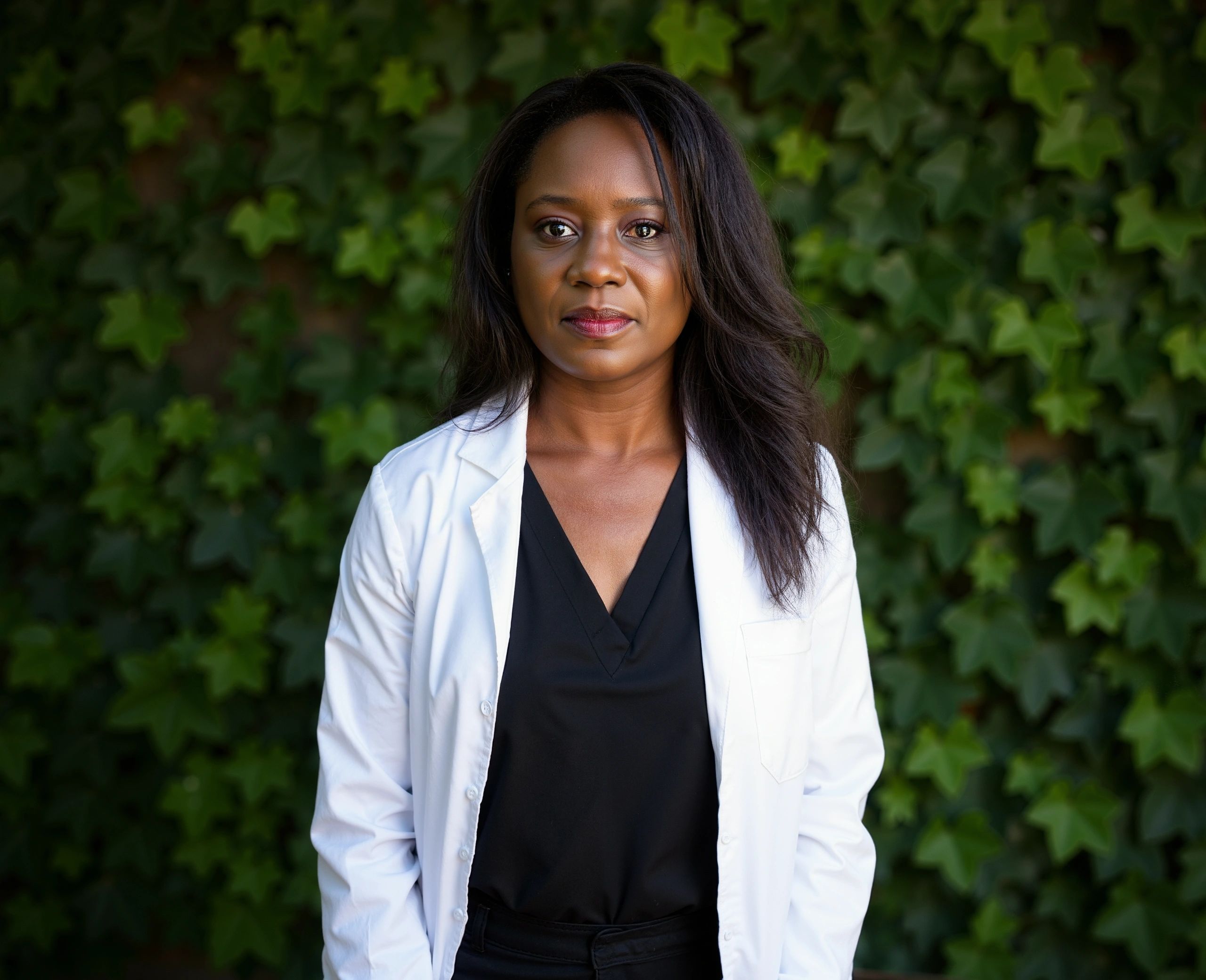 Confident female doctor in a white coat standing against a leafy background.