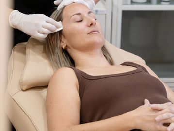 A client lays in the bed in clinic preparing for treatment while Cass cleans her face with a wipe