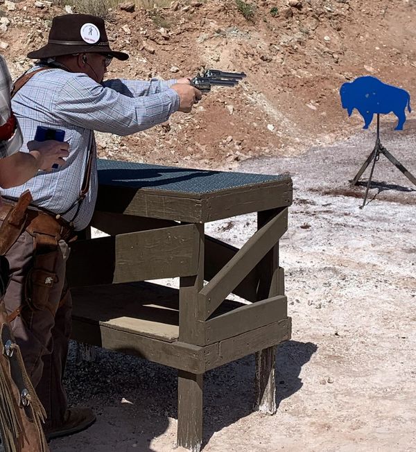 Man in cowboy attire shooting at a blue buffalo target outdoors.