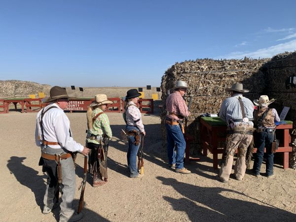 People dressed in cowboy attire stand in line with rifles at a shooting range.