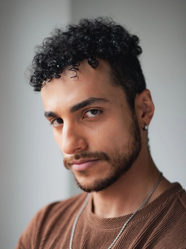 Confident young man with curly hair and a beard, wearing a brown shirt and silver chain.