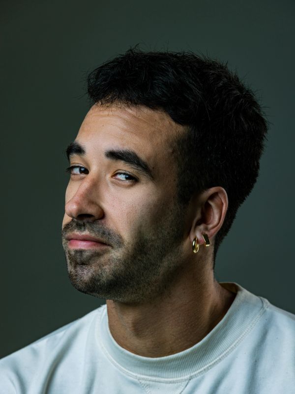Man with dark hair and gold earrings wearing a white shirt smirking.