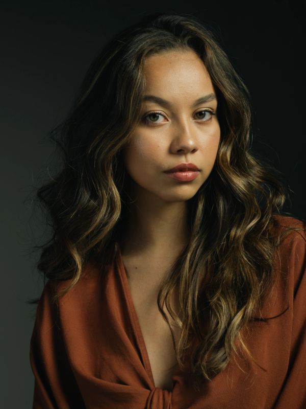 Portrait of a young woman with wavy hair in a brown top against a dark background.