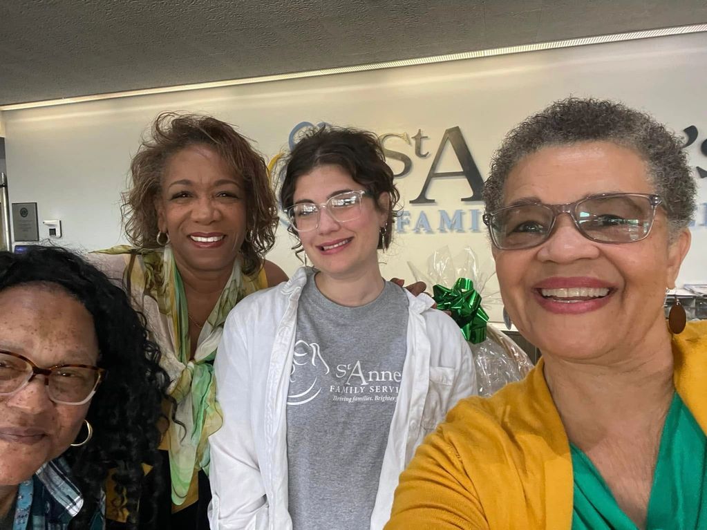 Four smiling women take a group selfie indoors.