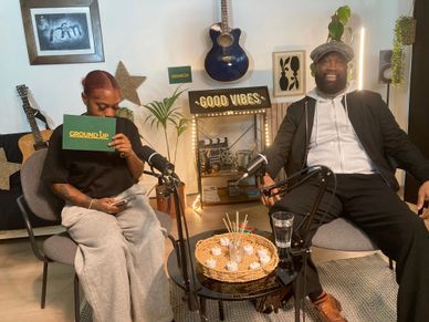 Two people smiling and chatting in a cozy podcast setup with guitars and warm decor.