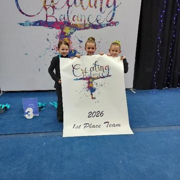 Three young gymnasts hold a banner for the 2026 1st Place Team.
