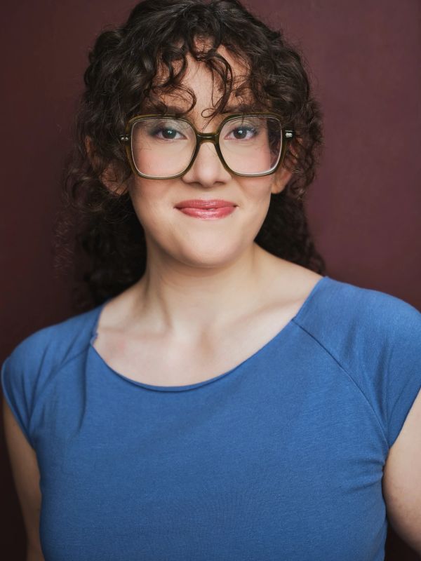 Woman with curly hair and glasses smiles warmly in blue top.