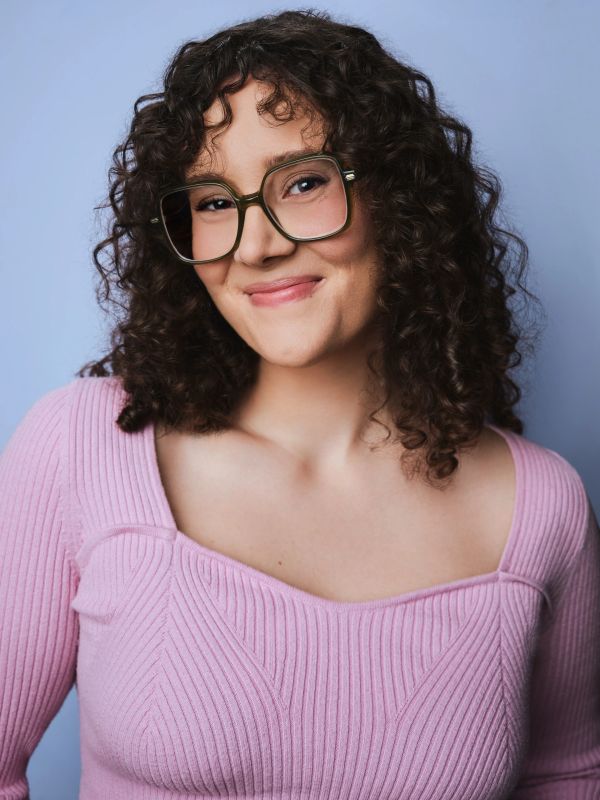 Smiling woman with curly hair and glasses wearing a pink sweater.