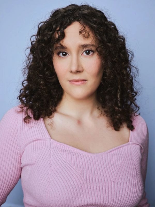 Woman with curly hair wearing a pink sweater against a blue background.