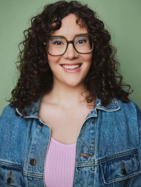 Smiling woman with curly hair wearing glasses and a denim jacket.