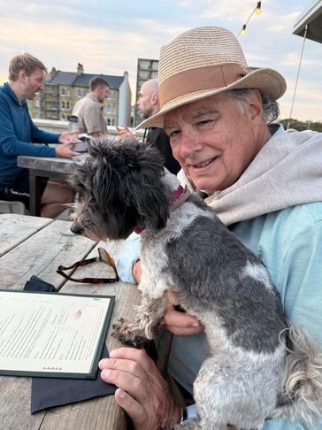 Man in a hat holding a small black and white dog at an outdoor table.