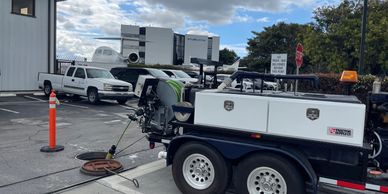 Utility trailer with hose reel near an open manhole in a parking lot with airplanes.