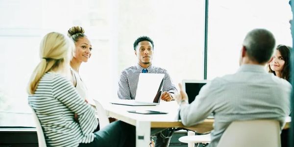 A diverse group of professionals in a meeting around a table with laptops and documents.