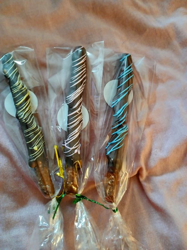 Three chocolate-covered pretzel rods with colorful drizzle, individually wrapped in clear plastic.