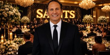 Man in a suit smiling at a formal event with 'SAINTS' sign in background.
