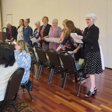 A group of women standing and holding papers in a room with wooden floors.