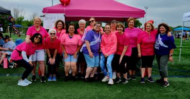 Group of women in pink shirts smiling under a pink tent at an outdoor event.