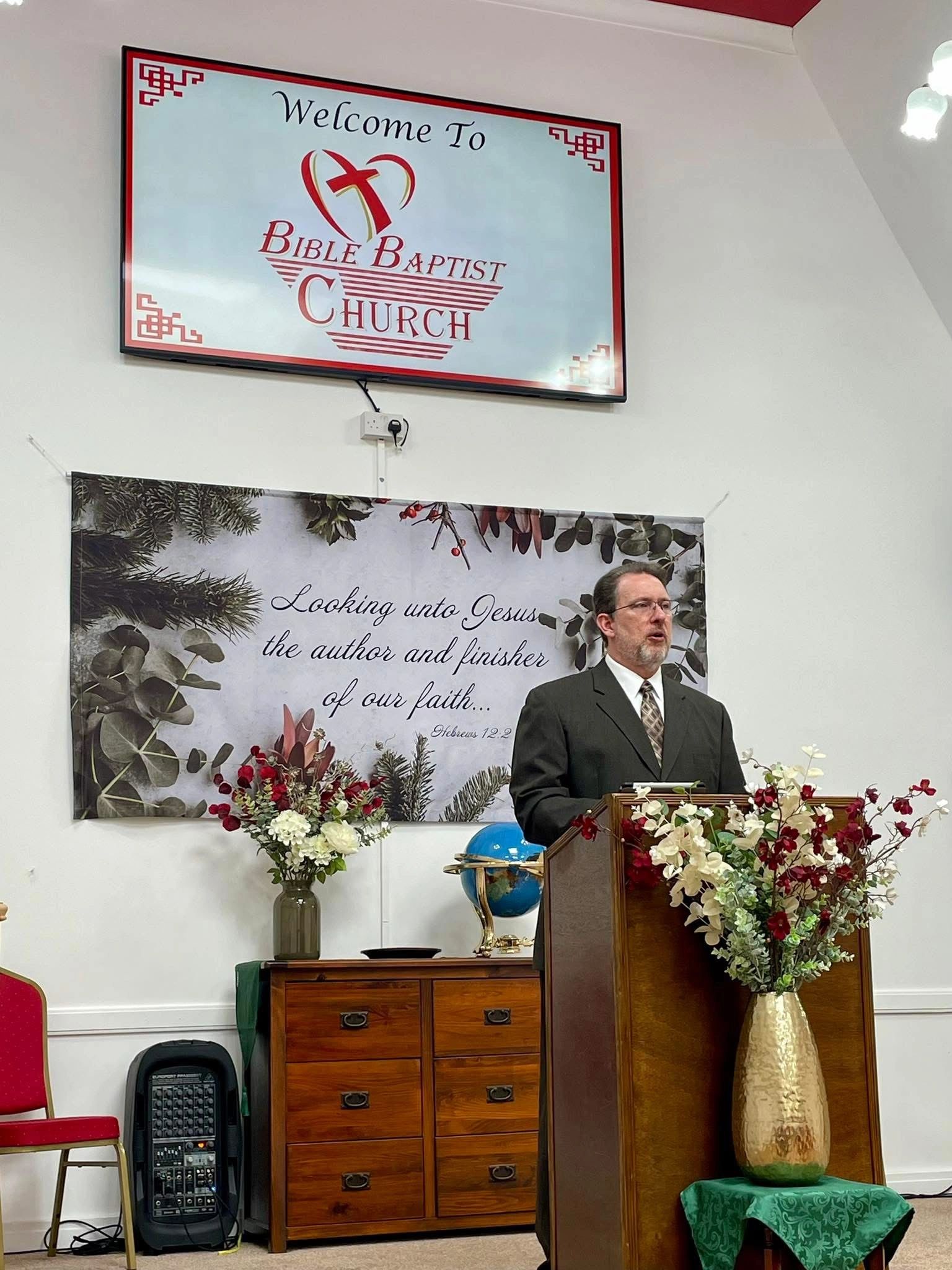 Man speaking at Bible Baptist Church podium with floral decorations.