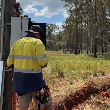 Worker in high-visibility clothing inspecting outdoor electrical equipment in a rural area.