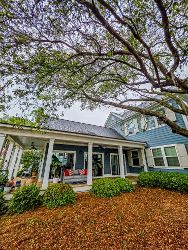 Cozy porch with seating under a large tree beside a blue house.