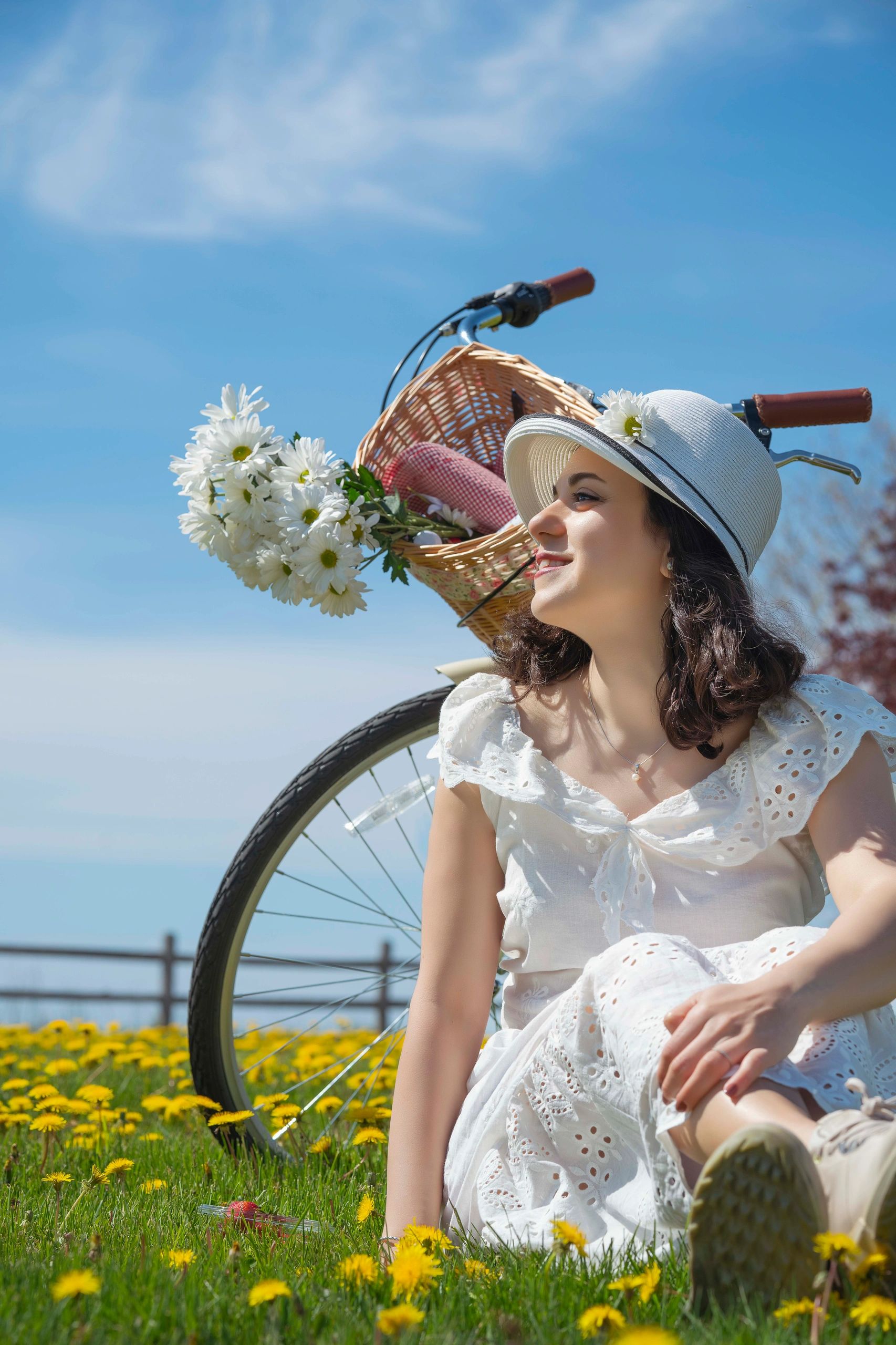 Young woman in white dress and hat sitting in a flower field beside a bicycle with daisies. Dreamy o
