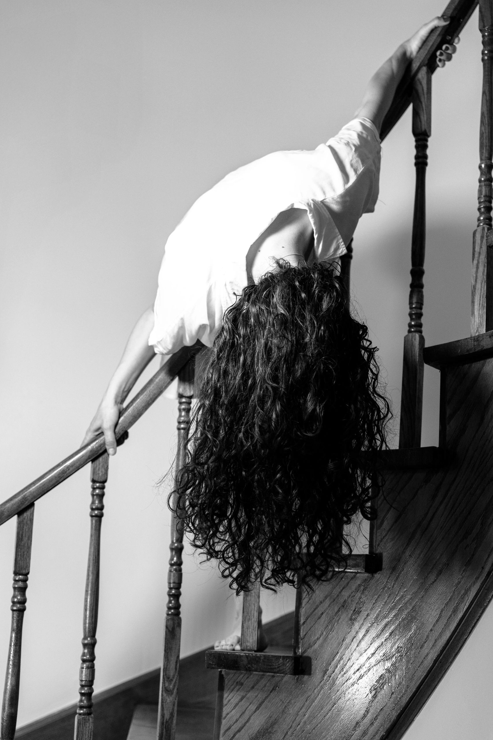 A person with long curly hair bends backward over a wooden staircase railing.Elegant personal portra