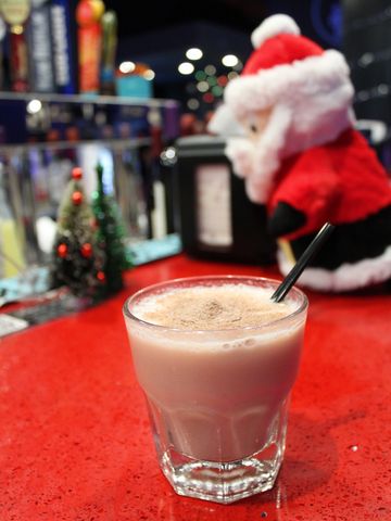 A glass of frothy beverage on a red table with a blurred Santa decoration in the background.