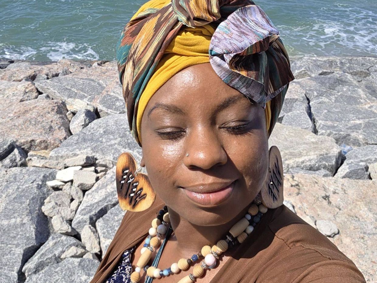 Woman with a colorful headwrap and large earrings smiles by the rocky shoreline.