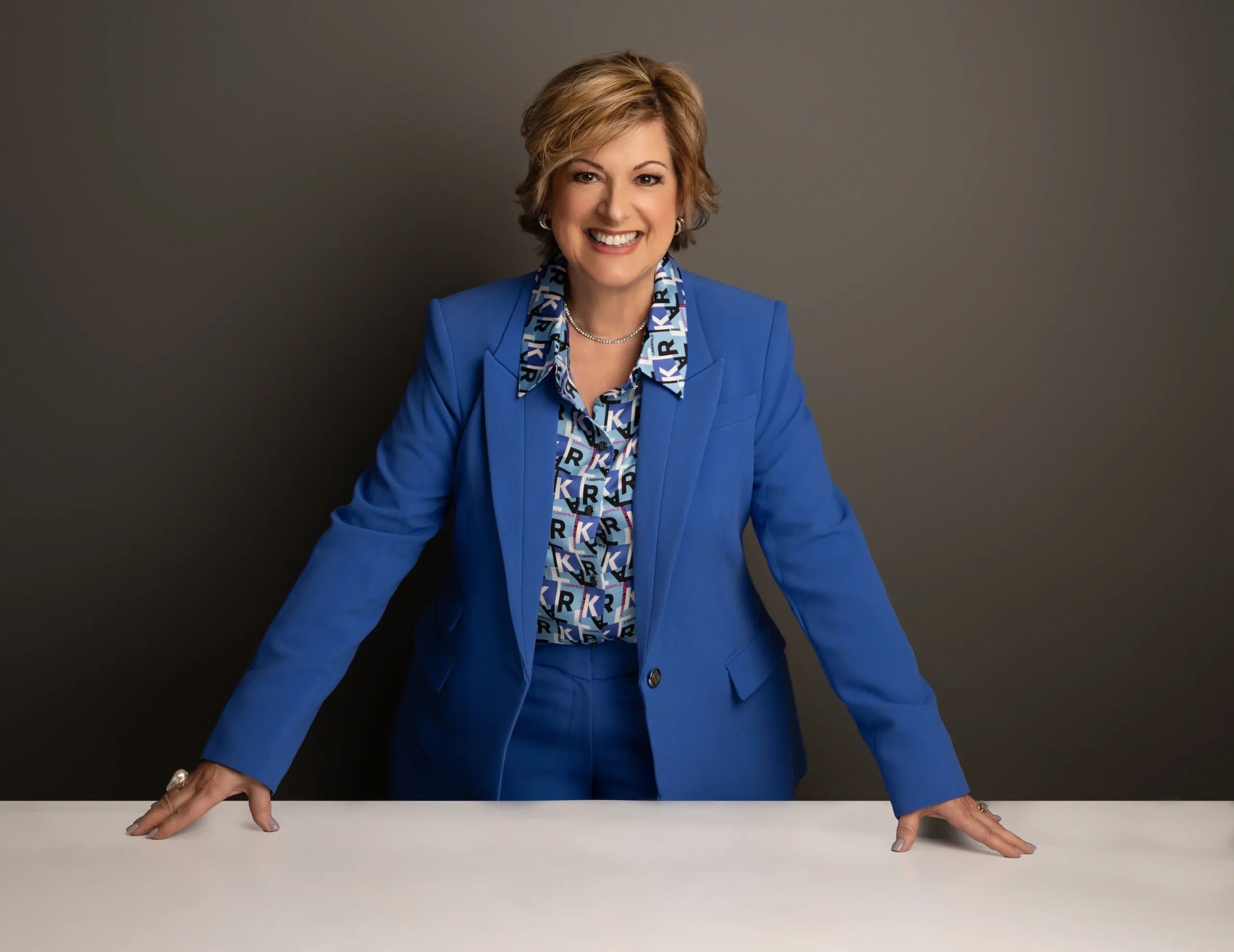 Confident woman in a bright blue suit smiles warmly, leaning on a white table.