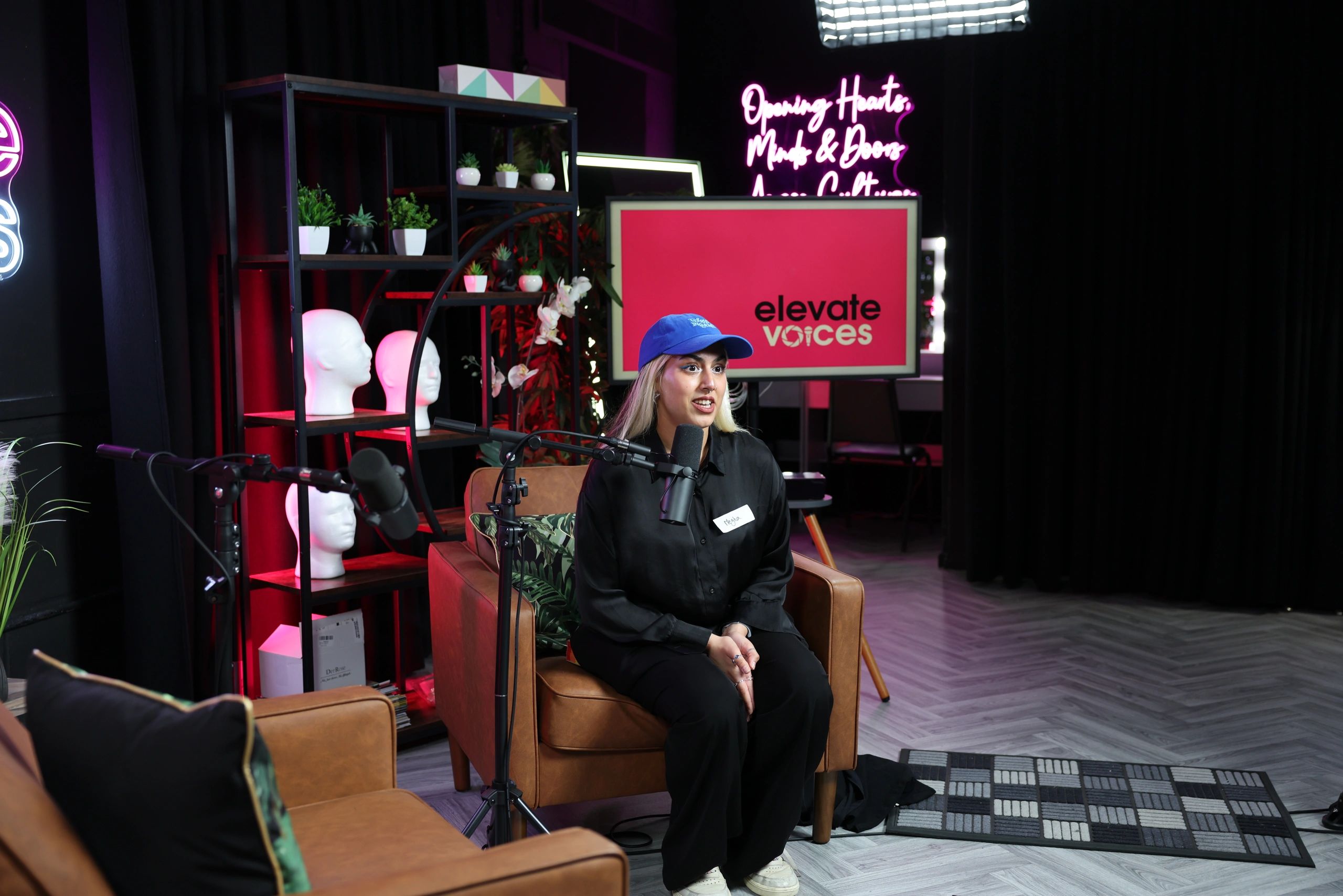 A woman wearing a blue cap speaks into a microphone in a cozy, decorated recording studio.